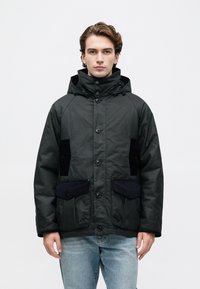 Tânăr îmbrăcat într-o jachetă parka neagră cu glugă și buzunare cu nasturi, purtând blugi, stând în fața unui fundal alb simplu.