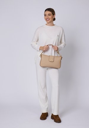 Beige läderhandväska med en strukturerad design, guldfärgade detaljer och ett avtagbart rem, hålls av en person i vitt mysigt klädd.