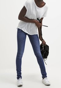 Camiseta blanca de manga corta, jeans skinny azules y zapatillas blancas. Sosteniendo un bolso negro con un detalle de cadena plateada. Atuendo simple y casual.