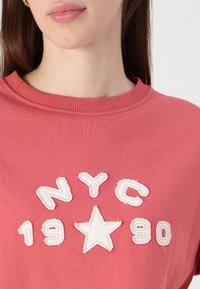 Sudadera rosa con texto bordado en blanco "NYC 1990" y diseño de estrella. Presenta un cuello redondo acanalado, textura suave y corte casual.