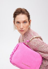 Modella con gli occhi blu e orecchini lancia uno sguardo intenso mentre tiene in mano una borsa a tracolla rosa intrecciata, indossando un maglione rosa di tessuto strutturato.