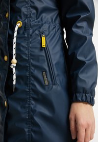 Námořnická voděodolná bunda s žebrovanými manžetami, kapucí se stahovací šňůrkou, žlutým zipem a bočními kapsami. Obsahuje logo značky na dolní části předního dílu.