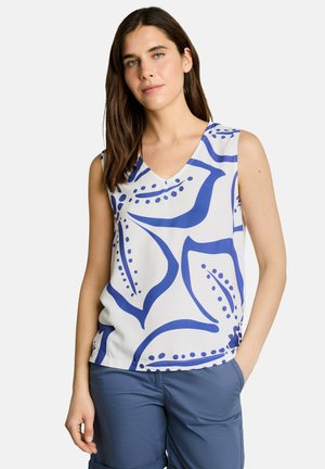 Femme portant un haut blanc sans manches avec un grand imprimé floral abstrait bleu et un short bleu, debout avec une main dans la poche.