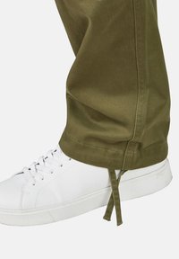 Olijfgroene broeken met een gladde textuur en een verstelbaar trekkoord aan de onderkant, gecombineerd met witte sneakers met een gestructureerde zool.