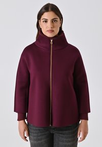 Vrouw in een hooggesloten pruimenjack met een gouden rits, geribbelde kraag en boorden, gecombineerd met donkergrijze jeans,staand tegen een witte achtergrond.