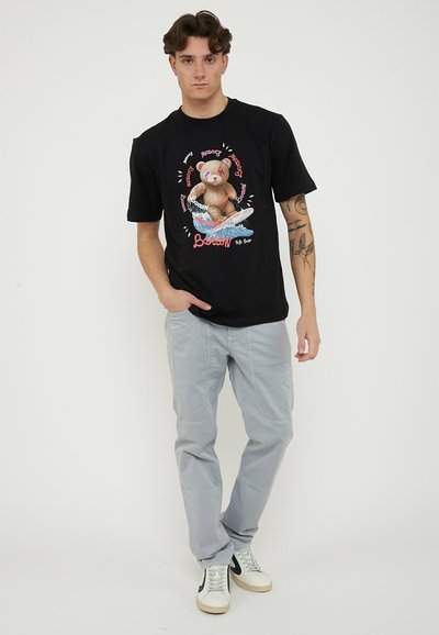 Young man with tattoos wearing black T-shirt featuring surfing teddy bear graphic, light grey pants, and white sneakers against plain background.