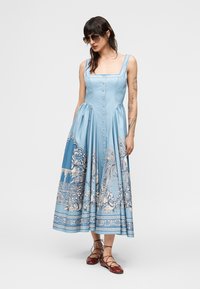Kobieta ubrana w jasnoniebieską sukienkę bez rękawów z guzikami z przodu oraz szczegółowym białym kwiatowym i architektonicznym wzorem, brązowe płaskie buty na sznurowadła oraz okulary przeciwsłoneczne.