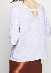 Top in lino a righe blu chiaro con maniche a tre quarti e dettaglio a chiave sul retro, abbinato a una gonna brillante di colore ruggine.