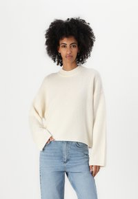 Kremowy, oversize'owy sweter z prążkowanym dekoltem i szerokimi rękawami, wykonany z miękkiego, teksturowanego materiału. Połączony z jasnoniebieskimi dżinsami.