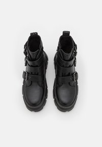 Μαύρες μπότες μέχρι τον αστράγαλο από υφασμάτινο δέρμα με πολλές μαύρες ζώνες και πόρπες. Στρογγυλή μύτη, χοντρή σόλα και λαβή στο πίσω μέρος.