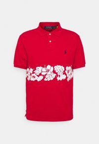 Röd pikétröja med ett vitt blommönster och bladmönster över bröstet, med en diskret marinblå logotyp på vänster sida.