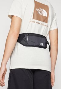 Musta vyölaukku, jossa on verkkoyksityiskohtia ja vetoketjukiinnitys, ja siinä on "The North Face" -logo. Käytetään vaaleanvärisen t-paidan päällä.