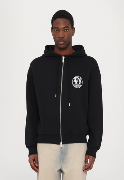 Black zip-up hoodie made of cotton with a large front pocket, drawstrings, and a round logo patch on the left chest.