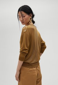 Maglione marrone con polsini e orlo a costine, dotato di fori metallici sulle spalle. Il modello indossa grandi orecchini geometrici, in posa di spalle.