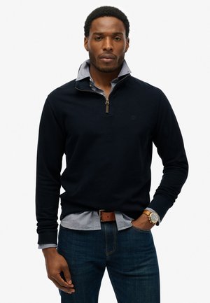 Marinefarbener Pullover mit Viertelreißverschluss, über einem hellblauen Hemd mit Kragen getragen. Verfügt über ein dezentes Logo und gerippte Bündchen.