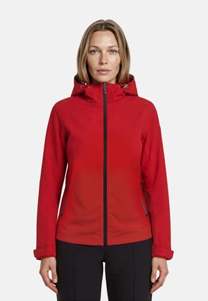 Femme portant une veste à capuche rouge, fermée jusqu'au cou, avec un pantalon noir, debout de face sur un fond blanc uni.