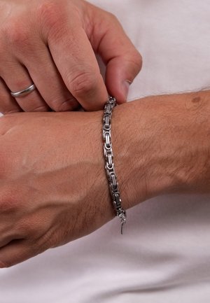Personne portant une bague en argent en train de fermer un bracelet chaîne épais en argent autour d'un poignet sur fond blanc.