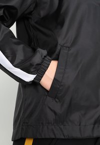 Giacca a vento nera con righe decorative bianche, con polsini elasticizzati e tasche laterali. Materiale liscio e leggero con finitura opaca.