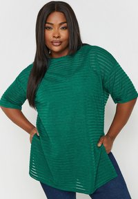 Grön texturerad topp med ribbat mönster, korta ärmar och lös passform. Materialet verkar mjukt med en lätt lyster. Kombinerad med mörka jeans.