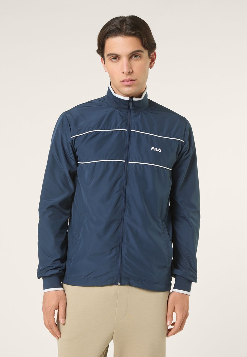 Nuori mies pukeutuneena laivastonsiniseen vetoketjulliseen takkiin, jossa on valkoiset koristeraidat ja FILA-logo, seisomassa yksinkertaisen vaalean taustan edessä.