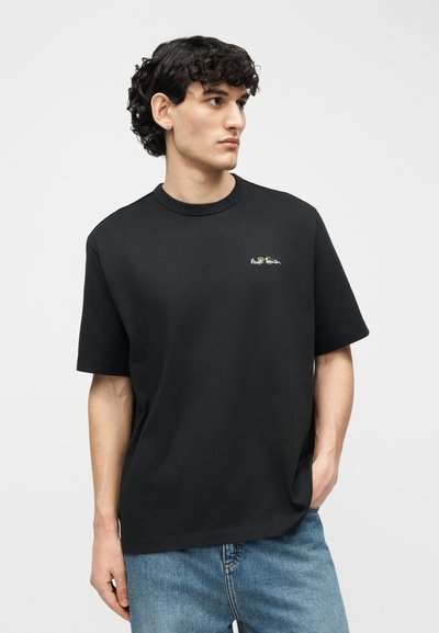 Νεαρός άνδρας με σγουρά σκουρόχρωμα μαλλιά, φοράει μαύρο κοντομάνικο μπλουζάκι με μικρό λογότυπο στο στήθος και μπλε τζιν, κοιτάζοντας προς τα δεξιά.