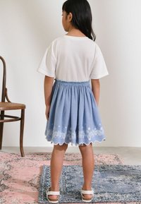 Weißes Kurzarm-T-Shirt, kombiniert mit einem hellblauen Rock mit einer gewellten Säume und weißen Blumenstickereien. Einfaches Design und leichtes Material.