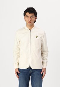 Lette cremejakke med stående krave, frontlynlås, ribbede manchetter og et lille gult logo på venstre bryst. Båret med blå jeans.