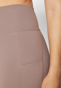 Gros plan sur un legging taille haute mauve avec des détails de coutures visibles et une poche latérale, montrant une partie d'un bras et du torse.