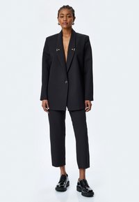 Zwarte op maat gemaakte blazer met scherpe revers en gouden hardware accenten, gecombineerd met zwarte broek tot de enkel en glanzende plateau schoenen.