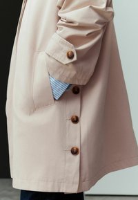 Bras dans un manteau beige avec de gros boutons marron et un poignet de chemise bleu rayé visible depuis la poche.
