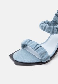 Ανοικτό πέδιλο από τζιν σε γαλάζιο χρώμα με λουριά με ρυτίδες και χαμηλό μαύρο τετράγωνο τακούνι σε λευκό φόντο.