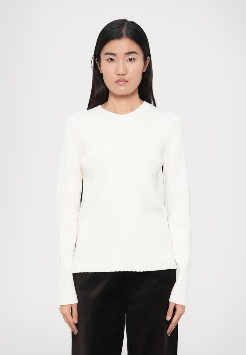 Suéter blanco acanalado con mangas largas y escote redondeado, combinado con pantalones oscuros de pierna ancha. Textura suave, ajuste estructurado.