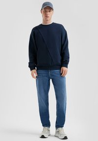 Felpa navy con design asimmetrico, polsini a costine, abbinata a jeans azzurri e sneakers grigie. Il cappello è in denim azzurro.
