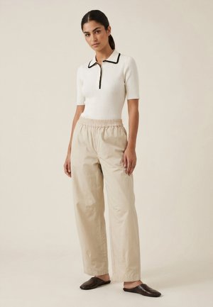 Femme debout portant un polo blanc à manches courtes côtelé, un pantalon beige à jambes larges et des chaussures marron foncé sans lacets.
