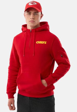 Uomo con felpa rossa dei Kansas City Chiefs con cappuccio e scritta gialla, e cappellino rosso abbinato con logo della squadra, mano in tasca, su sfondo uniforme.