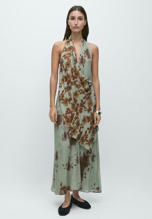 Femme portant une robe verte fluide longue jusqu’aux chevilles avec des motifs floraux marron et des chaussures plates noires, debout devant un fond clair uni.