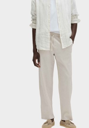 Mężczyzna model noszący beżowe spodnie na sznurku, jasnokremową koszulę w paski z podwiniętymi rękawami, białą podkoszulkę oraz beżowe mokasyny z frędzlami.