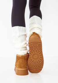 Bottines en suede marron avec une semelle en caoutchouc texturée. Des chauffe-jambes blancs côtelés contrastent avec des leggings noirs.