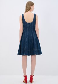 Farkkutunika dengan rok melebar, leher belakang berbentuk baskom, jahitan vertikal, dan sandal hak merah. Tunik ini berwarna biru tua dengan jahitan yang terlihat.