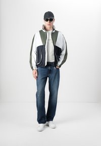 Fargeblokkert jakke i grønn, hvit og marineblå med høy krage, kombinert med lyseblå vide jeans og hvite joggesko. Svart caps og solbriller.