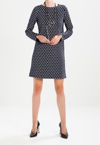 Marineblaues Kleid mit langen Ärmeln und einem Rautenmuster. A-Linie Silhouette, aus weichem Stoff gefertigt. Kombiniert mit schwarzen High-Heels und geschichteten Halsketten.
