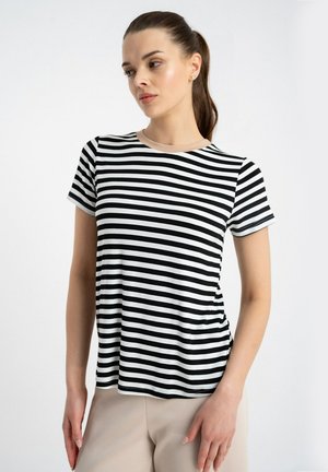 Kobieta z brązowymi włosami związanymi w kucyk, ubraną w czarno-biały poziomo paskowany krótki rękaw i beżowe spodnie, patrzy w lewo.