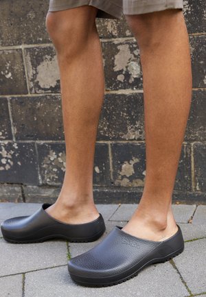 Zoccoli neri in gomma con superficie texturizzata, schienale aperto e suola spessa, posizionati su una superficie pavimentata con gambe visibili.