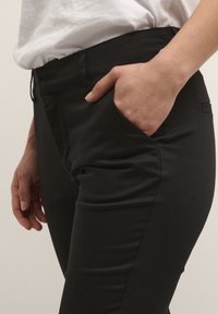 Pantalon noir à coupe ajustée, présentant une texture lisse, des poches avant et une ceinture. La main repose dans la poche pour le contexte.