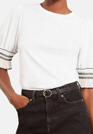Femme portant un chemisier blanc avec des poignets de manches brodés noirs et un jean noir taille haute avec une ceinture noire et une boucle ronde dorée.