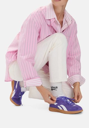 Fioletowe buty sportowe z białymi akcentami, wykonane z zamszu i siateczki, w połączeniu z białymi spodniami i różową koszulą w paski, osoba wiążąca sznurowadła.