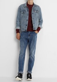 Světle modrá džínová bunda s dvěma kapsami na prsou a knoflíky. Pod ní vínová mikina s kulatým výstřihem. Světle modré džíny. Černé tenisky.