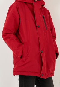 Punainen eristetty takki hupulla, jossa vetoketjullinen tasku ja napillinen suljenta, valmistettu sileästä kankaasta mattapintaisella viimeistelyllä.