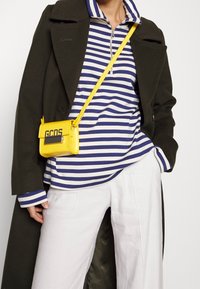 Gul läderaxelväska med lock och logotyp, tillsammans med en blå och vit randig långärmad skjorta och vita vida byxor.