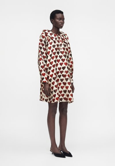 Γυναίκα που φοράει ένα φαρδύ φόρεμα μέχρι το γόνατο με κόκκινα και καφέ σχέδια καρδιών, μακριά μανίκια, μεγάλο γιακά και μαύρα χαμηλά τακούνια με μυτερή μύτη.
